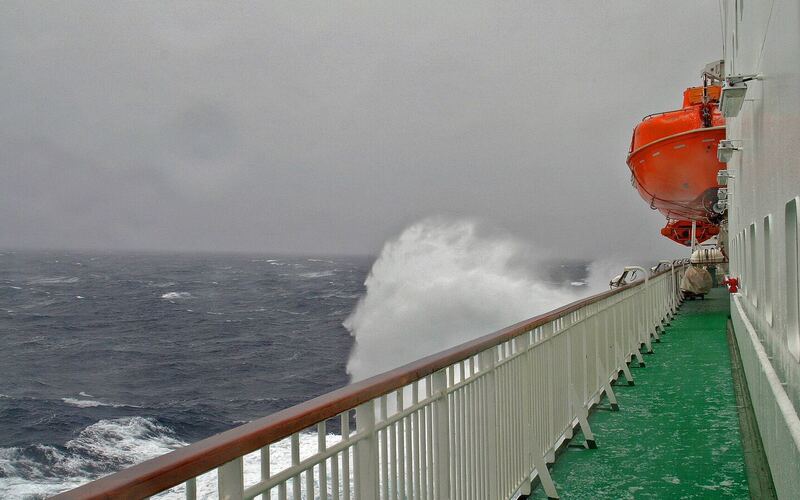 Een golf slaat schuimend over de reling aan de zijkant van een schip op de grijze, woeste zee van de Drake Passage