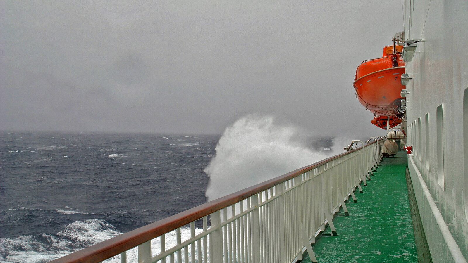 Een golf slaat schuimend over de reling aan de zijkant van een schip op de grijze, woeste zee van de Drake Passage
