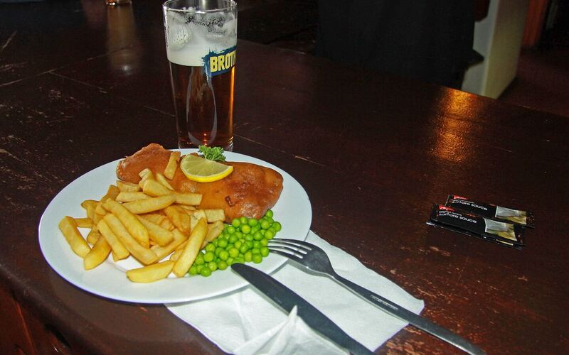Een bord fish and chips met erwten en een schijfje citroen naast een glas bier op een houten pubtafel in Stanley