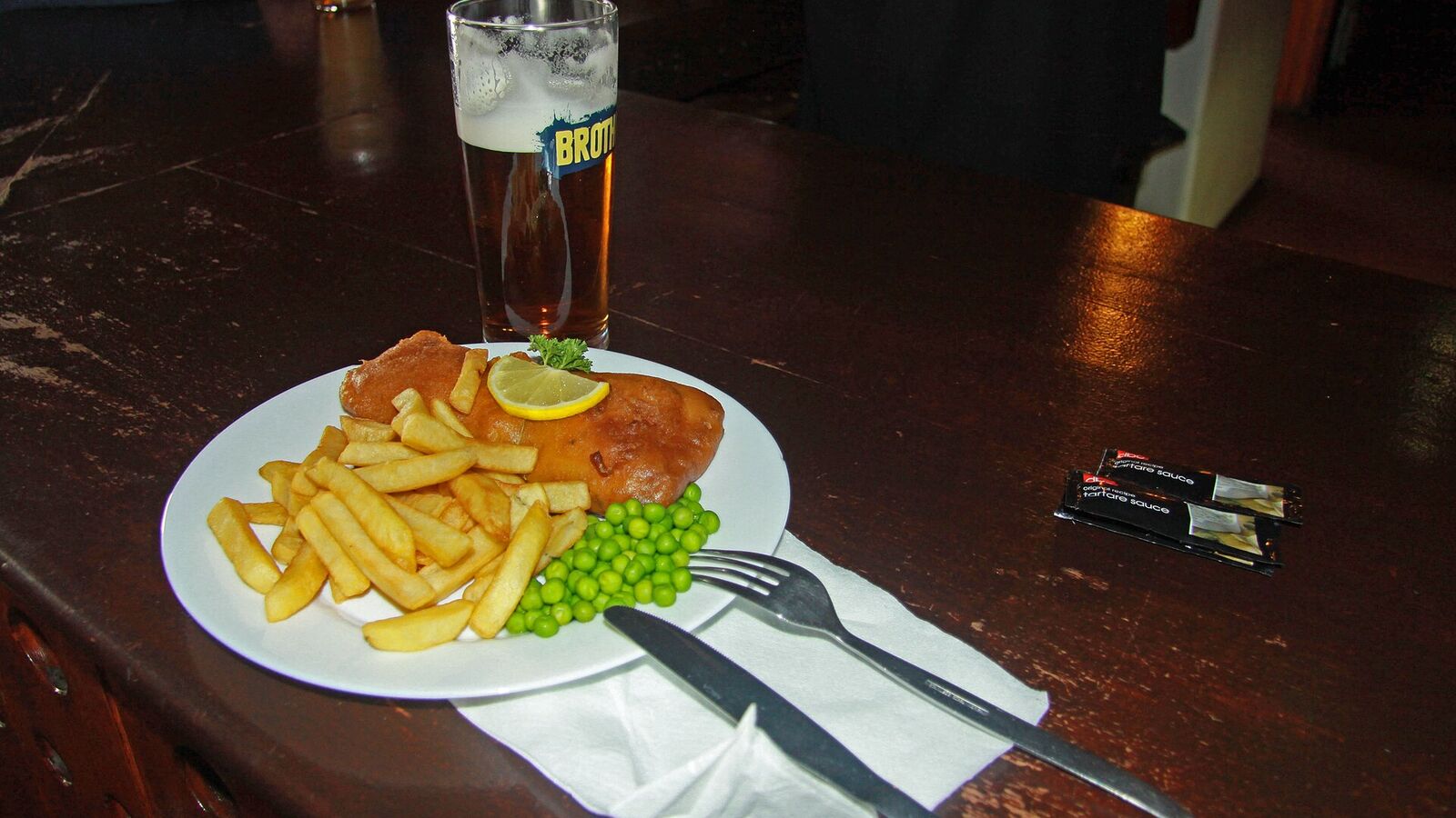 Een bord fish and chips met erwten en een schijfje citroen naast een glas bier op een houten pubtafel in Stanley