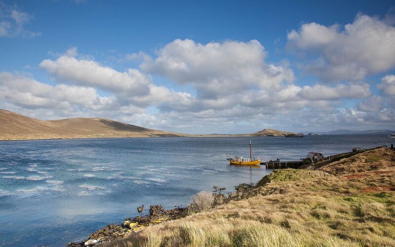 Winderige baai bij Carcass Island met wit schuimende golven, geel grasland op de oever en een kleine boot aan de steiger