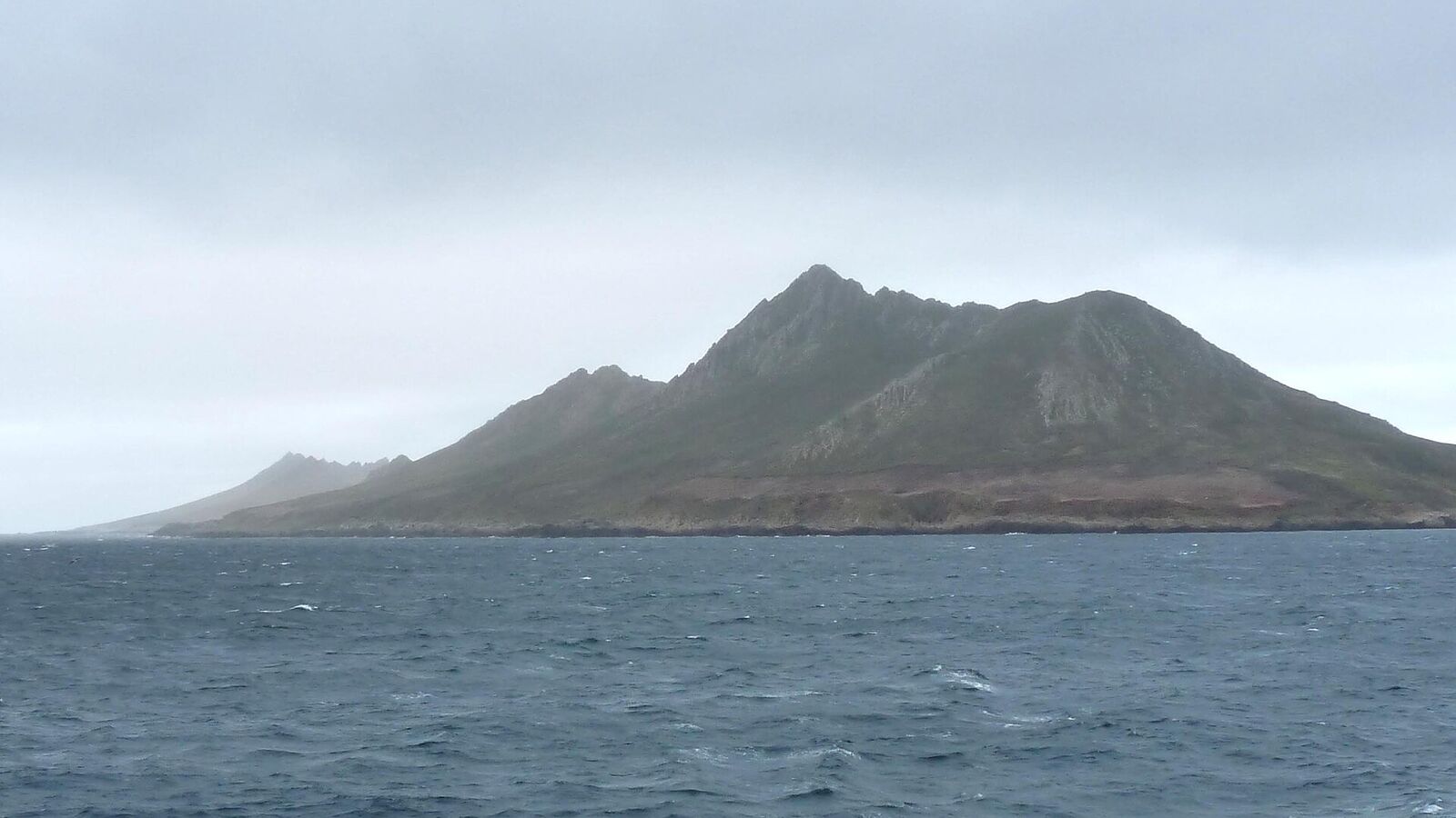 Het bergachtige eiland Grand Jason rijst op uit een grijsblauwe zee onder een bewolkte lucht