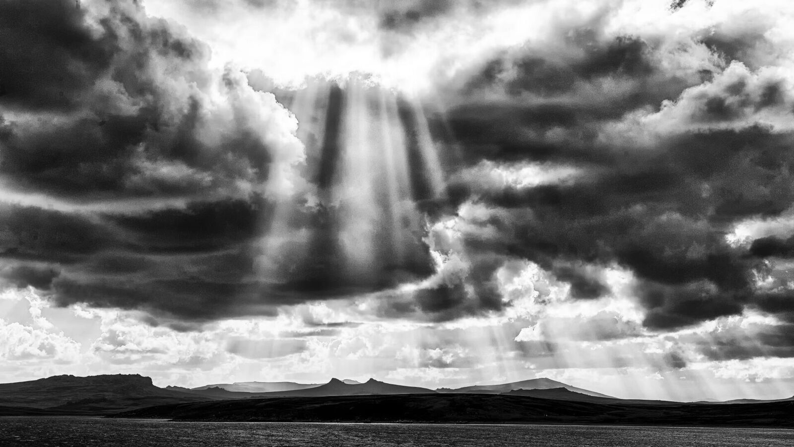 Zwart-witfoto van zonnestralen die door donkere wolken breken boven een laag landschap op de Falklands